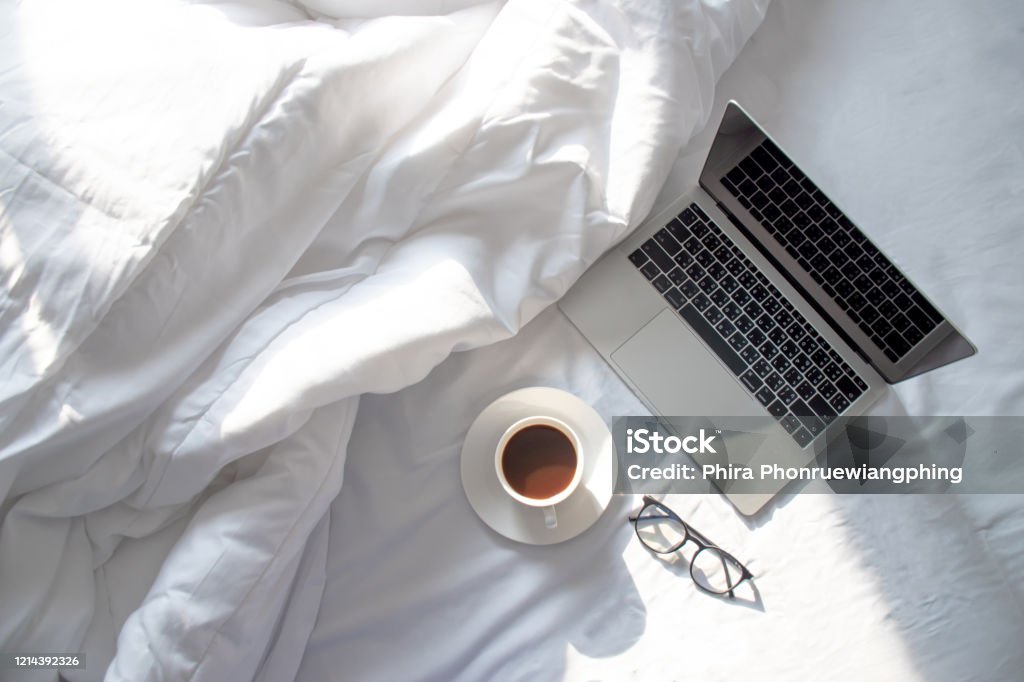 Top view of laptop and hot coffee in bed with soft sunlight in the morning. Bed - Furniture Stock Photo