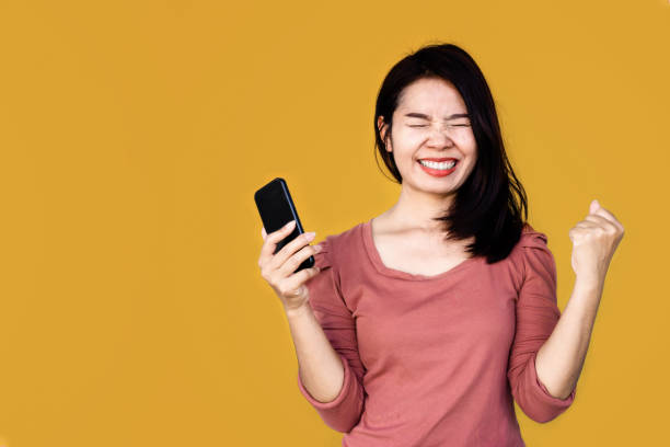 fröhliche fröhliche asiatische frau erhalten gute nachrichten hand halten smartphone über gelben hintergrund - smiling single object photography orange stock-fotos und bilder