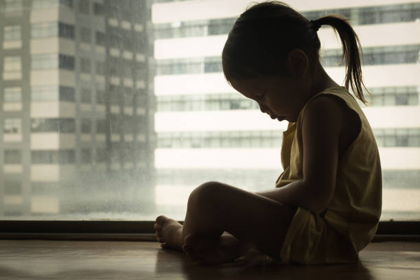 Sad child cryng alone. Depression. A litte girl sitting next to a window with her head down in sadness. Feeling depressed and hurt. face down stock pictures, royalty-free photos & images