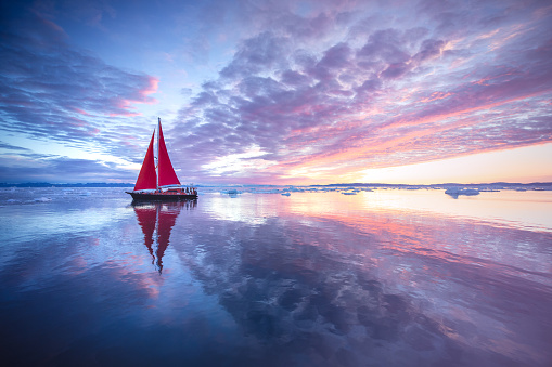 Red sail boat cruising among ice bergs in Greenland.