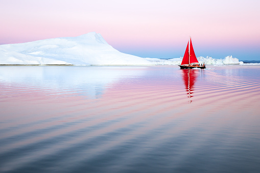 Sail boat with red sails cruising among ice bergs during midnight sun season. Disko Bay, Greenland.