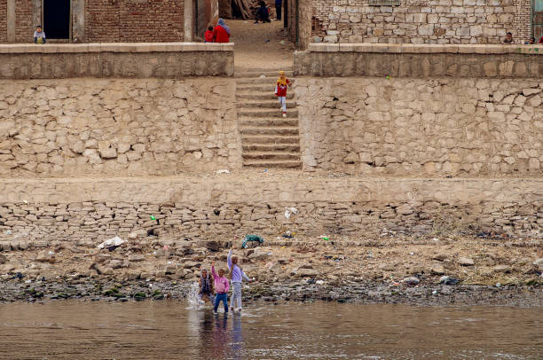 daily life in egypt. children playing in the nile, singing and cheering to the tourists passing by on the daily cruises, with their small town in the background - desert egyptian culture village town imagens e fotografias de stock