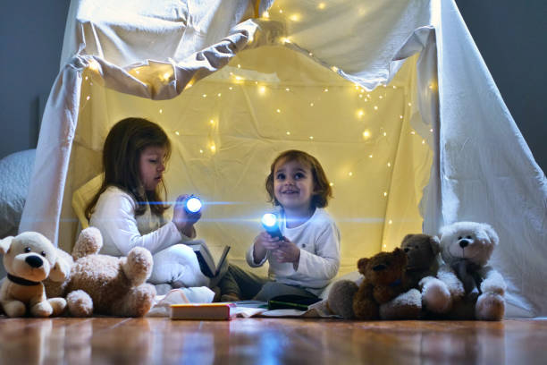 deux petites filles jouent à la maison dans la soirée pour construire une tente de camping pour lire des livres avec une lampe de poche et dormir à l’intérieur - wigwam photos et images de collection