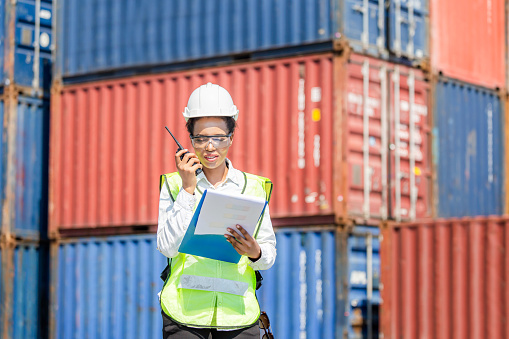 Woman Foreman in hardhat and safety vest holding holding clipboard checklist and talks on two-way radio control loading containers box from cargo