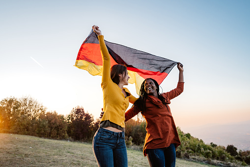 Two young pretty multi-ethnic women holding German flag on meadow.