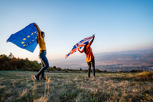 Two young pretty multi-ethnic women holding European Union and British flag on meadow. Brexit.