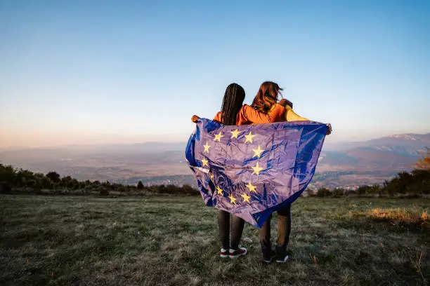 Two young pretty multi-ethnic women holding European Union flag on meadow.