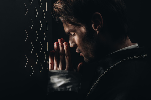joven sacerdote católico rezando con los ojos cerrados cerca de la rejilla confesional en la oscuridad con rayos de luz photo