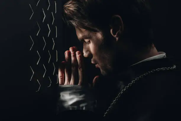 Photo of young catholic priest praying with closed eyes near confessional grille in dark with rays of light