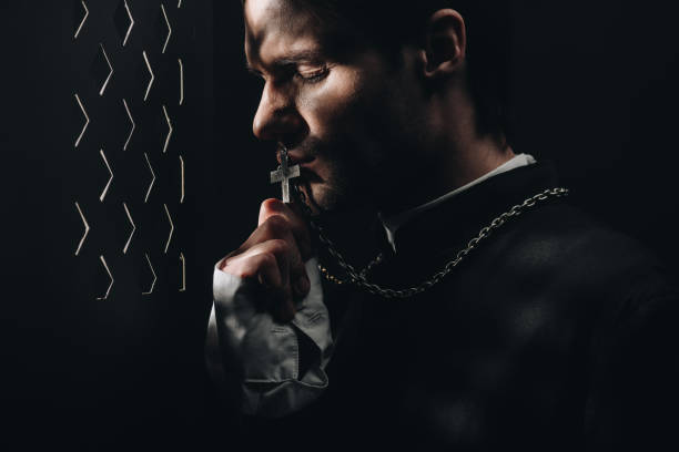 joven sacerdote católico tenso besando cruz en su collar en la oscuridad cerca de la rejilla confesional con rayos de luz - confession booth fotografías e imágenes de stock