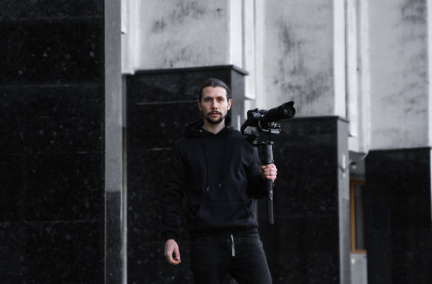 istock Young Professional videographer holding professional camera on 3-axis gimbal stabilizer. Pro equipment helps to make high quality video without shaking. Cameraman wearing black hoodie making a videos. 1214462123
