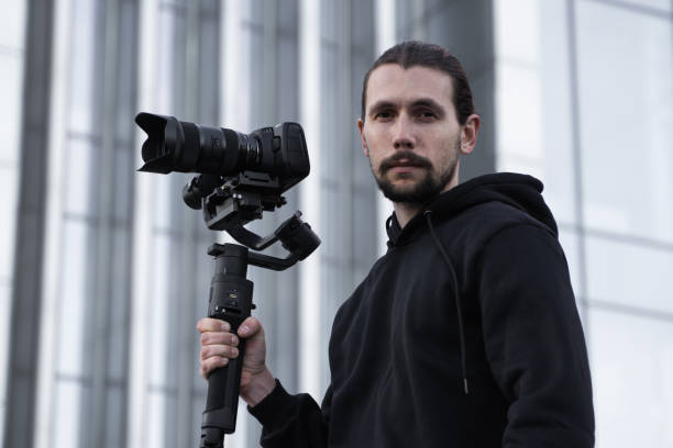 istock Young Professional videographer holding professional camera on 3-axis gimbal stabilizer. Pro equipment helps to make high quality video without shaking. Cameraman wearing white hoodie making a videos. 1214462203