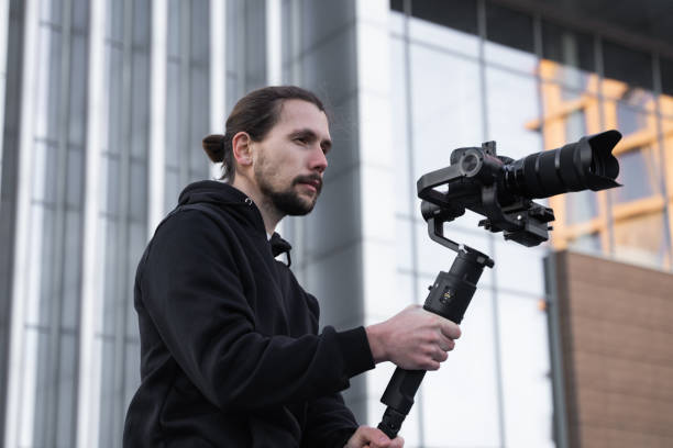istock Young Professional videographer holding professional camera on 3-axis gimbal stabilizer. Pro equipment helps to make high quality video without shaking. Cameraman wearing white hoodie making a videos. 1214462289