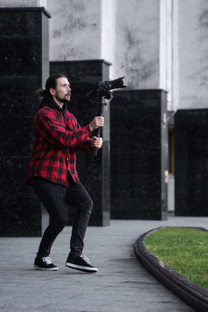 istock Young Professional videographer holding professional camera on 3-axis gimbal stabilizer. Pro equipment helps to make high quality video without shaking. Cameraman wearing red shirt making a videos. 1214462417