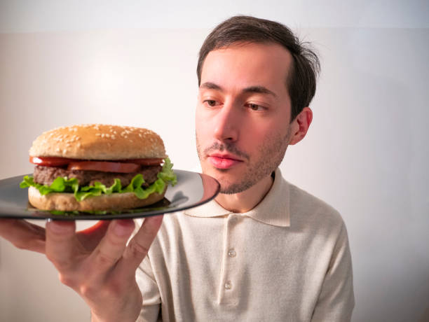 istock Handsome young man holding, looking and examining tasty hamburger for hexamination 1214462456