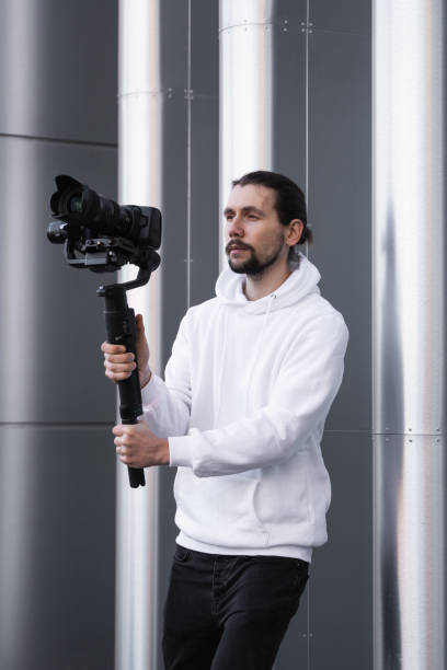 istock Young Professional videographer holding professional camera on 3-axis gimbal stabilizer. Pro equipment helps to make high quality video without shaking. Cameraman wearing white hoodie making a videos. 1214462512