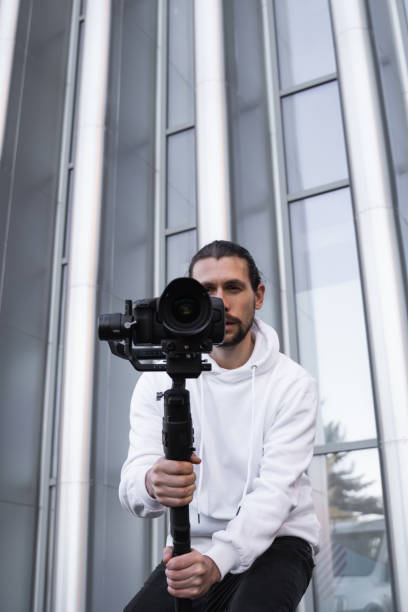 istock Young Professional videographer holding professional camera on 3-axis gimbal stabilizer. Pro equipment helps to make high quality video without shaking. Cameraman wearing white hoodie making a videos. 1214463055