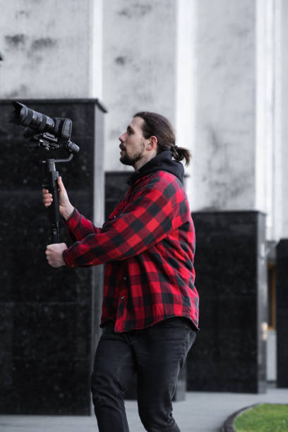 istock Young Professional videographer holding professional camera on 3-axis gimbal stabilizer. Pro equipment helps to make high quality video without shaking. Cameraman wearing red shirt making a videos. 1214463127