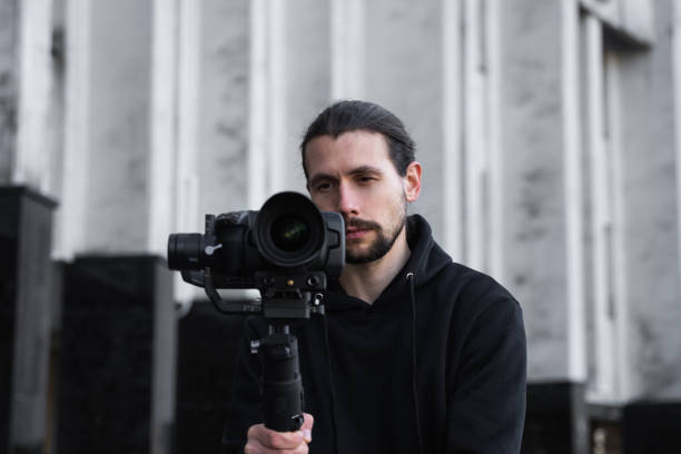 istock Young Professional videographer holding professional camera on 3-axis gimbal stabilizer. Pro equipment helps to make high quality video without shaking. Cameraman wearing black hoodie making a videos. 1214463224