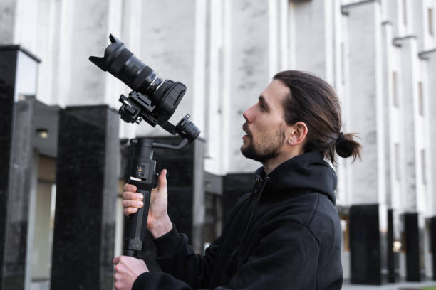 istock Young Professional videographer holding professional camera on 3-axis gimbal stabilizer. Pro equipment helps to make high quality video without shaking. Cameraman wearing black hoodie making a videos. 1214463349