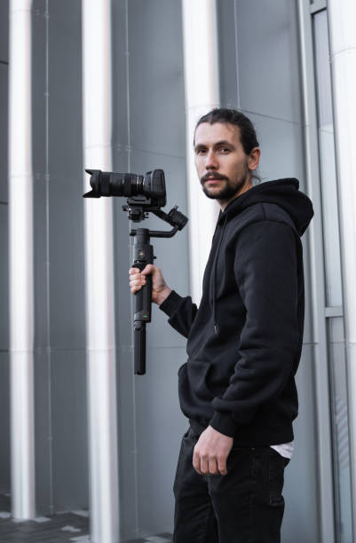 istock Young Professional videographer holding professional camera on 3-axis gimbal stabilizer. Pro equipment helps to make high quality video without shaking. Cameraman wearing white hoodie making a videos. 1214463462