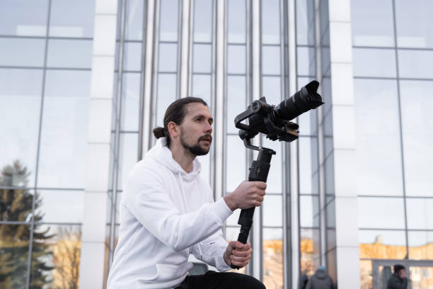 istock Young Professional videographer holding professional camera on 3-axis gimbal stabilizer. Pro equipment helps to make high quality video without shaking. Cameraman wearing white hoodie making a videos. 1214463677