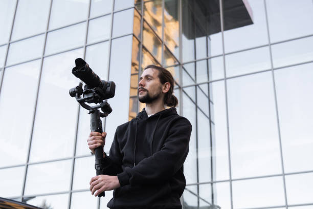 Young Professional videographer holding professional camera on 3-axis gimbal stabilizer. Pro equipment helps to make high quality video without shaking. Cameraman wearing white hoodie making a videos.