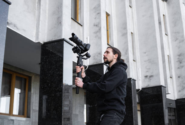 istock Young Professional videographer holding professional camera on 3-axis gimbal stabilizer. Pro equipment helps to make high quality video without shaking. Cameraman wearing black hoodie making a videos. 1214463871