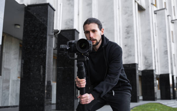 istock Young Professional videographer holding professional camera on 3-axis gimbal stabilizer. Pro equipment helps to make high quality video without shaking. Cameraman wearing black hoodie making a videos. 1214464507