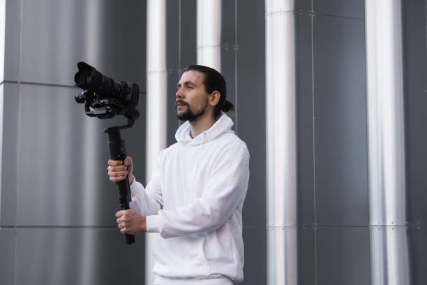 istock Young Professional videographer holding professional camera on 3-axis gimbal stabilizer. Pro equipment helps to make high quality video without shaking. Cameraman wearing white hoodie making a videos. 1214465218