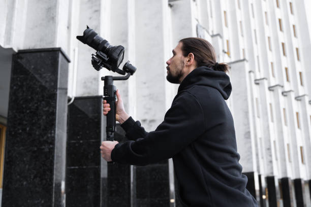 istock Young Professional videographer holding professional camera on 3-axis gimbal stabilizer. Pro equipment helps to make high quality video without shaking. Cameraman wearing black hoodie making a videos. 1214465302