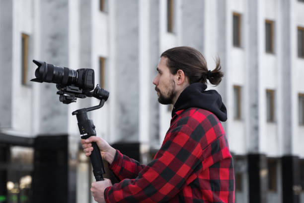 istock Young Professional videographer holding professional camera on 3-axis gimbal stabilizer. Pro equipment helps to make high quality video without shaking. Cameraman wearing red shirt making a videos. 1214465414