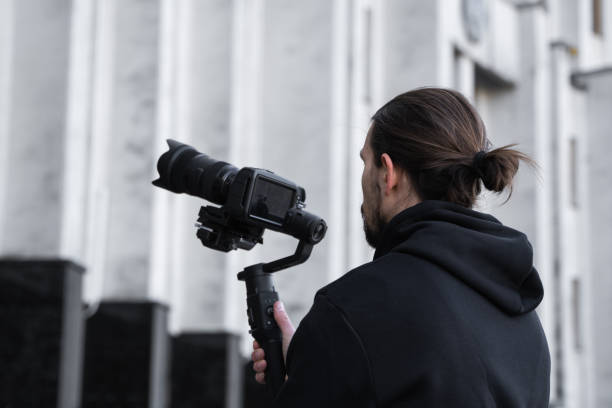 istock Young Professional videographer holding professional camera on 3-axis gimbal stabilizer. Pro equipment helps to make high quality video without shaking. Cameraman wearing black hoodie making a videos. 1214465494
