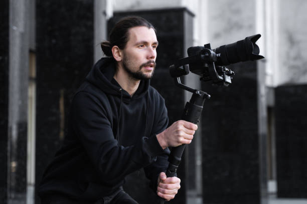 istock Young Professional videographer holding professional camera on 3-axis gimbal stabilizer. Pro equipment helps to make high quality video without shaking. Cameraman wearing black hoodie making a videos. 1214465581