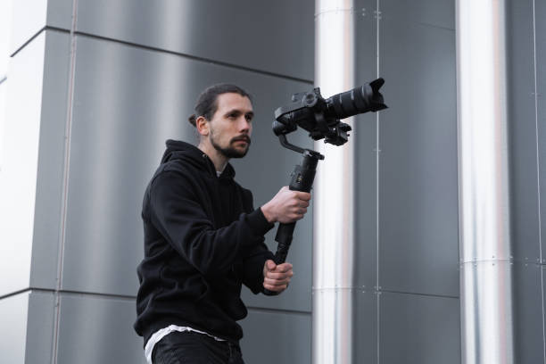 istock Young Professional videographer holding professional camera on 3-axis gimbal stabilizer. Pro equipment helps to make high quality video without shaking. Cameraman wearing white hoodie making a videos. 1214465926