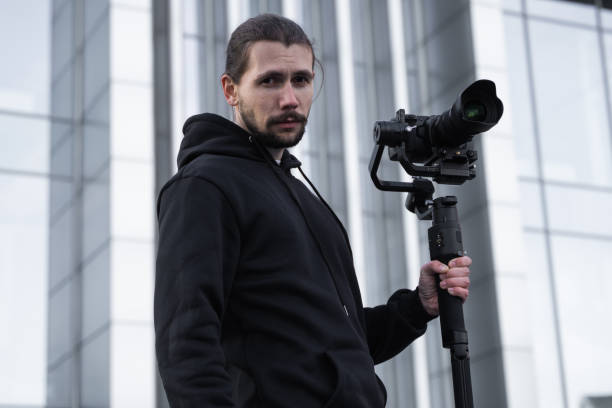 istock Young Professional videographer holding professional camera on 3-axis gimbal stabilizer. Pro equipment helps to make high quality video without shaking. Cameraman wearing white hoodie making a videos. 1214466003