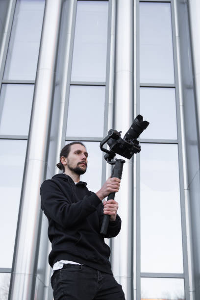 istock Young Professional videographer holding professional camera on 3-axis gimbal stabilizer. Pro equipment helps to make high quality video without shaking. Cameraman wearing white hoodie making a videos. 1214466099