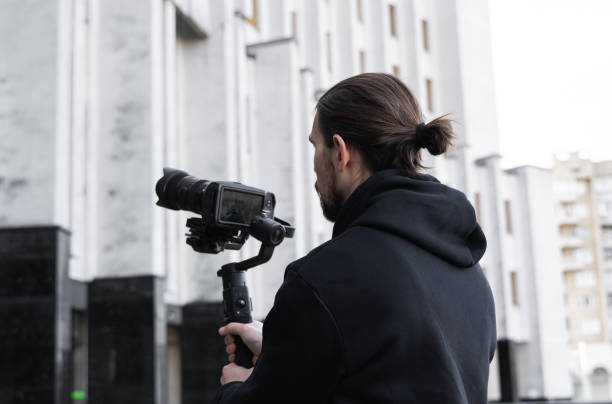 istock Young Professional videographer holding professional camera on 3-axis gimbal stabilizer. Pro equipment helps to make high quality video without shaking. Cameraman wearing black hoodie making a videos. 1214466177