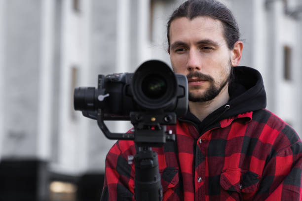 istock Young Professional videographer holding professional camera on 3-axis gimbal stabilizer. Pro equipment helps to make high quality video without shaking. Cameraman wearing red shirt making a videos. 1214467239