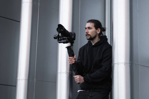 istock Young Professional videographer holding professional camera on 3-axis gimbal stabilizer. Pro equipment helps to make high quality video without shaking. Cameraman wearing white hoodie making a videos. 1214467618