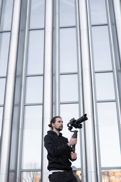istock Young Professional videographer holding professional camera on 3-axis gimbal stabilizer. Pro equipment helps to make high quality video without shaking. Cameraman wearing white hoodie making a videos. 1214467745