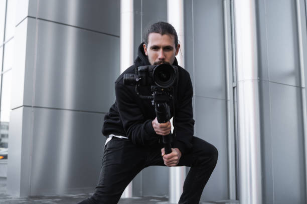 Young Professional videographer holding professional camera on 3-axis gimbal stabilizer. Pro equipment helps to make high quality video without shaking. Cameraman wearing white hoodie making a videos.