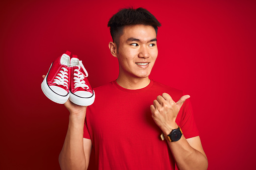 Young asian chinese man holding casual sneakers standing over isolated red background pointing and showing with thumb up to the side with happy face smiling