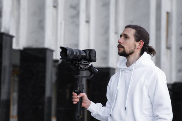 istock Young Professional videographer holding professional camera on 3-axis gimbal stabilizer. Pro equipment helps to make high quality video without shaking. Cameraman wearing white hoodie making a videos. 1214469122