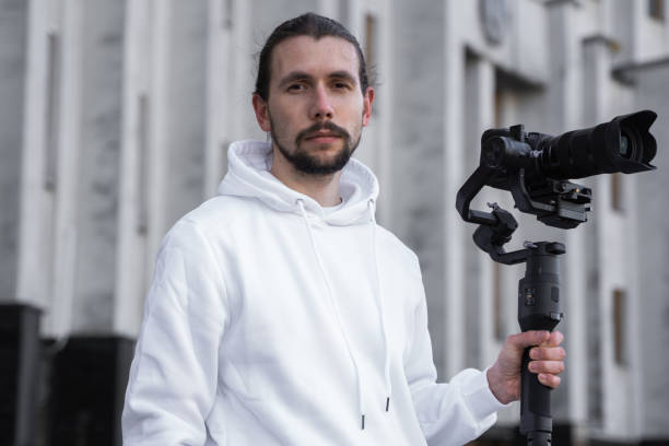 istock Young Professional videographer holding professional camera on 3-axis gimbal stabilizer. Pro equipment helps to make high quality video without shaking. Cameraman wearing white hoodie making a videos. 1214469418