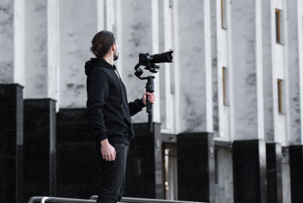 istock Young Professional videographer holding professional camera on 3-axis gimbal stabilizer. Pro equipment helps to make high quality video without shaking. Cameraman wearing black hoodie making a videos. 1214469639
