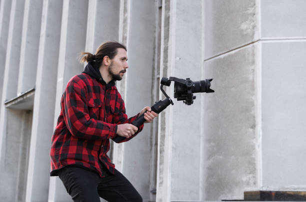 istock Young Professional videographer holding professional camera on 3-axis gimbal stabilizer. Pro equipment helps to make high quality video without shaking. Cameraman wearing red shirt making a videos. 1214469745