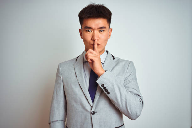 homme d’affaires chinois asiatique utilisant le costume et la cravate restant au-dessus du fond jaune isolé demandant à être calme avec le doigt sur des lèvres. silence et concept secret. - suit necktie close up gray photos et images de collection