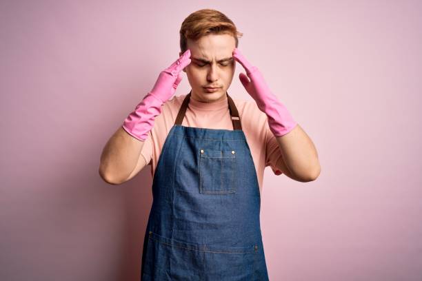istock Young handsome redhead cleaner man doing housework wearing apron and gloves with hand on head for pain in head because stress. Suffering migraine. 1214473683