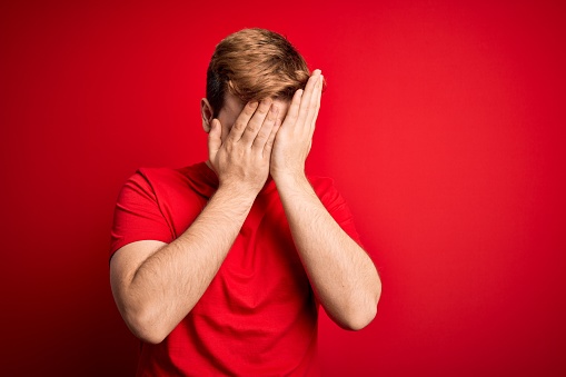 Young handsome redhead man wearing casual t-shirt over isolated red background with sad expression covering face with hands while crying. Depression concept.